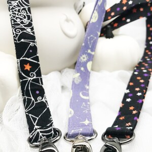 May include: Three Halloween-themed lanyards with metal swivel clasps. One is black with a white spiderweb design, another is lavender with celestial patterns, and the third is black with orange, purple, and white stars. The lanyards are displayed against a white background.