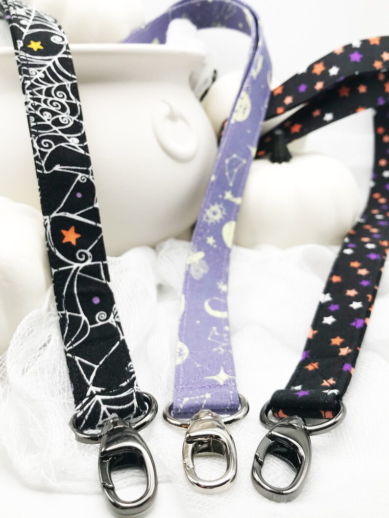 May include: Three Halloween-themed lanyards with metal swivel clasps. One is black with a white spiderweb design, another is lavender with celestial patterns, and the third is black with orange, purple, and white stars. The lanyards are displayed against a white background.