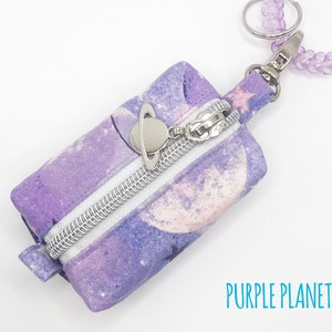 K&ouml;nnte beinhalten: Eine kleine, violette, mit einem Rei&szlig;verschluss versehene Tasche mit einem Galaxie-Druck und einem silbernen Anh&auml;nger. Die Tasche ist an einem lila Perlen-Schl&uuml;sselanh&auml;nger befestigt. Der Text "PURPLE PLANETS" ist auf einem wei&szlig;en Hintergrund unter der Tasche gedruckt.