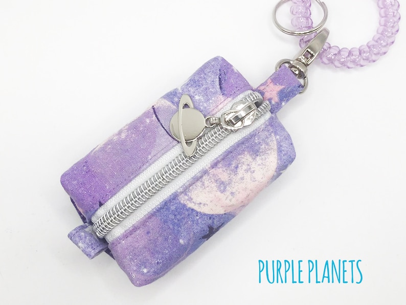 K&ouml;nnte beinhalten: Eine kleine, violette, mit einem Rei&szlig;verschluss versehene Tasche mit einem Galaxie-Druck und einem silbernen Anh&auml;nger. Die Tasche ist an einem lila Perlen-Schl&uuml;sselanh&auml;nger befestigt. Der Text "PURPLE PLANETS" ist auf einem wei&szlig;en Hintergrund unter der Tasche gedruckt.