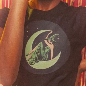 Smiling woman with an afro wearing a black graphic tee featuring a vintage-style green witch sitting on a crescent moon, holding a disco ball in front of shimmering pink fringe party decor. Perfect for retro witch fashion, magical party vibes, or Halloween aesthetic.