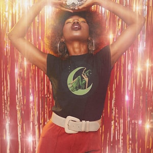 May include: A person wearing a black t-shirt with a green and white graphic of a witch on a crescent moon, paired with red pants and a white belt. A disco ball is held above the head. The background is a red tinsel curtain.