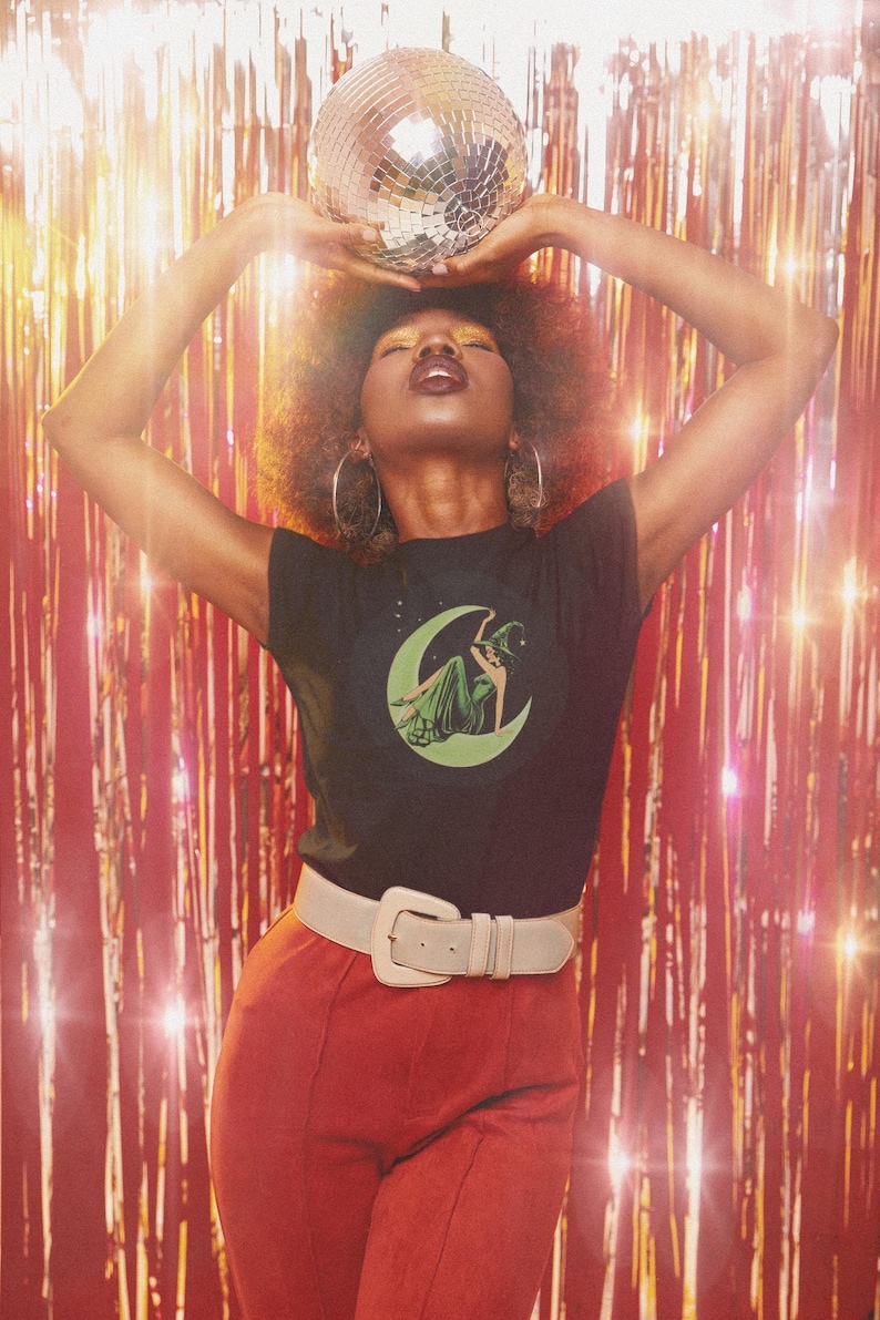 May include: A person wearing a black t-shirt with a green and white graphic of a witch on a crescent moon, paired with red pants and a white belt. A disco ball is held above the head. The background is a red tinsel curtain.