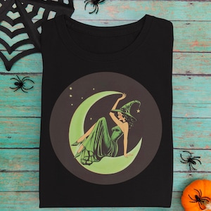 May include: Black t-shirt featuring a vintage-style illustration of a witch in a green dress and hat, seated on a crescent moon. The design is set within a dark brown circle, with stars in the background. Halloween-themed design.