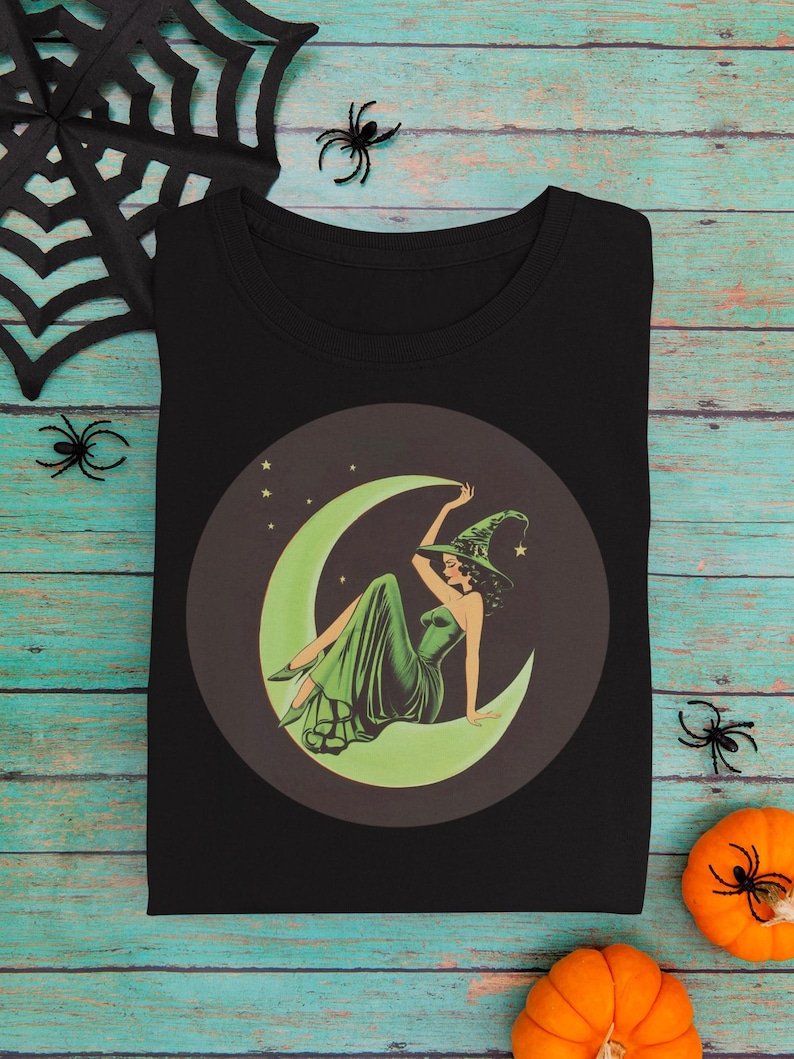 May include: Black t-shirt featuring a vintage-style illustration of a witch in a green dress and hat, seated on a crescent moon. The design is set within a dark brown circle, with stars in the background. Halloween-themed design.