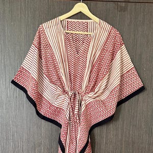 May include: A red and white patterned kaftan dress with a black trim. The dress features a V-neck, a drawstring waist, and a mix of vertical stripes and chevron patterns. The dress is hanging on a wooden hanger.