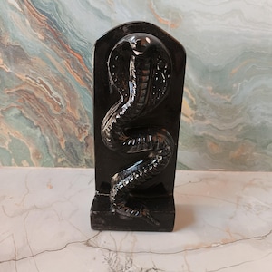 Könnte beinhalten: Eine schwarze, geschnitzte Kobraskulptur aus Stein. Die Kobra ist mit erhobener Haube dargestellt, ihr Körper krümmt sich nach unten. Die Skulptur steht vor einem marmorierten Hintergrund. Die Skulptur ist etwa 25 cm hoch.