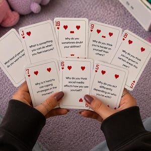 May include: A hand holds a deck of playing cards with red hearts and questions printed on them. The cards are white with red suit symbols and numbers. The questions include topics like self-doubt and social media's impact.