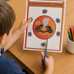 Op de afbeelding: Een kind knipt met een blauwe schaar een papieren knutselwerkje met een vulkaanthema uit. Het papier heeft een vulkaanillustratie met de tekst "VOLCANO GLOW" en de instructies: "Knip langs de zwarte lijn. Zorg ervoor dat je de lava niet aanraakt! Houd je duim op de vingerafdrukken als je het papier omdraait."