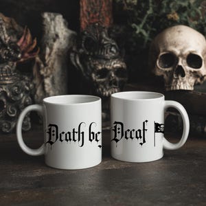 Peut inclure: Deux mugs en céramique blanche avec du texte gothique noir qui dit "Death be Decaf" et un drapeau pirate. Les mugs sont sur une surface sombre avec des crânes et d'autres décors gothiques en arrière-plan. Les mugs sont pour le café ou le thé.
