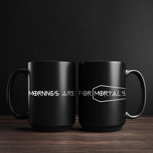 May include: Two black ceramic mugs with the text "Mornings are for Mortals" in white. One mug features the text, and the other has a coffin graphic with the same text. The mugs are on a wooden surface.