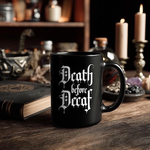 Peut inclure: Mug en céramique noire avec l'inscription blanche "Death before Decaf" en lettres gothiques. La tasse est posée sur une table en bois, avec des bougies et un livre en arrière-plan, créant une ambiance gothique.