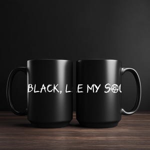 Peut inclure: Deux mugs noirs en céramique avec le texte blanc "BLACK, LIE MY SOUL" et un symbole de pentagramme. Les mugs sont posés sur une surface en bois sombre avec un fond noir.