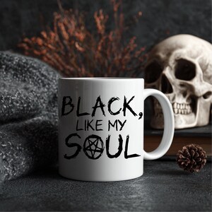 Peut inclure: Tasse en céramique blanche avec le texte "BLACK, LIKE MY SOUL" en lettres gothiques noires. La tasse présente un symbole de pentagramme. L'arrière-plan comprend un crâne, un article en tricot gris et des plantes séchées.