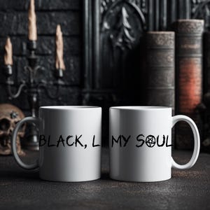 Peut inclure: Deux mugs en céramique blanche avec le texte noir "BLACK, L MY SOUL" et un symbole de pentagramme. Les mugs sont sur une surface sombre avec un fond gothique comprenant un crâne, des bougies et des livres.