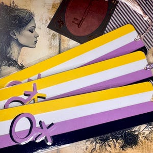 May include: Several non-binary pride flag bookmarks. Each bookmark has a yellow, white, purple, and black striped design with a silver non-binary symbol. The bookmarks are on a decorative background with tassels.
