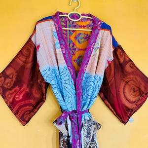 May include: A kimono-style robe featuring a patchwork design with various patterns and colors, including deep red sleeves with spiral motifs, a light blue section with circular patterns, and a bright orange and purple floral-patterned collar.