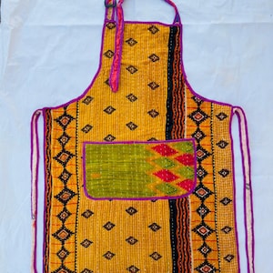 May include: A handmade apron with a yellow background and a green pocket. The apron features a geometric pattern in black, orange, and red. The edges are trimmed with purple fabric, and the ties are purple and white.