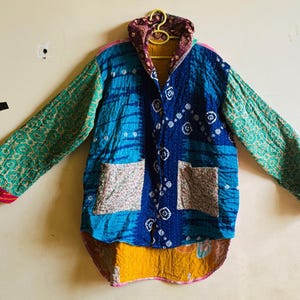 May include: A long-sleeve jacket featuring a patchwork design with blue, green, and yellow fabric panels. The garment includes a high collar, two front pockets with a floral pattern, and visible stitching throughout the quilted material.