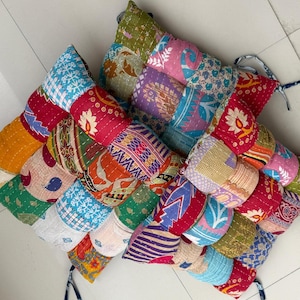 May include: A colorful, patchwork cushion with a woven design. The cushion is made of various fabrics in shades of red, blue, green, and purple. It has a quilted texture and decorative ties.