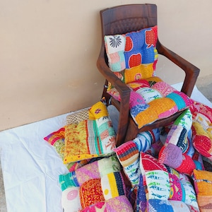May include: A wooden chair with colorful patchwork cushions. The cushions feature a variety of patterns and colors, including red, blue, yellow, and green. The chair is set against a neutral background.