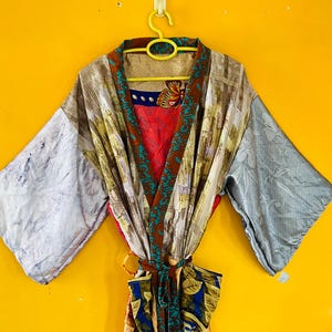 Pode incluir: Um roupão tipo quimono multicolorido com um design de patchwork que inclui vários padrões florais, de folhas e abstratos em tons de vermelho, azul, castanho e cinzento. A peça tem mangas largas, um cinto para atar e uma bainha contrastante.
