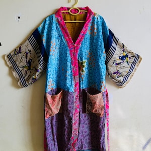 May include: A knee-length kimono robe featuring a patchwork design with various patterns in blue, purple, and pink. The garment has wide sleeves with bird illustrations, a bright pink front trim, two front pockets, and a matching fabric tie belt.