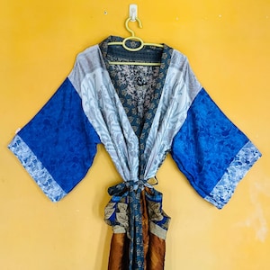 May include: A colorful kimono robe with a floral pattern. The robe features blue sleeves with a lace trim, a gray and white upper section, and a patterned tie. The robe is hanging on a yellow wall.