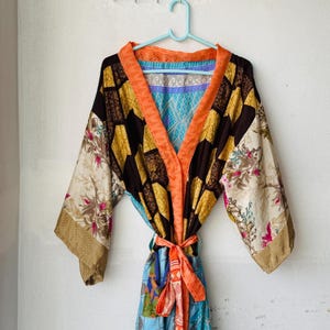 May include: A long-sleeved kimono robe featuring a patchwork design with panels of black, yellow, and cream fabric. The garment includes floral patterns, a bright orange collar and waist tie, and blue geometric accents on the inner lining and hem.