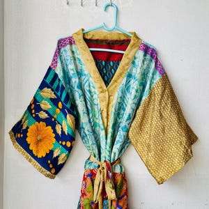 Puede incluir: Una bata estilo kimono con un patchwork de telas vibrantes. La manga izquierda muestra una gran flor amarilla sobre un fondo azul oscuro con hojas doradas, mientras que la manga derecha es dorada con un patrón geométrico.