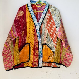 Op de afbeelding: Een patchwork jasje met diverse kleuren en patronen, waaronder rood, geel, oranje en bloemmotieven. De jas heeft twee zakken en een zwarte bies. Het hangt aan een gele hanger.