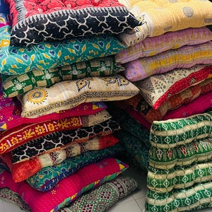 May include: A stack of square floor cushions featuring various colorful patterns, including red, yellow, green, and pink fabrics with intricate geometric, floral, and kantha-style stitching. The cushions are tufted and arranged in two piles.