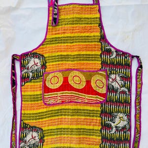 Puede incluir: Un delantal colorido, hecho a mano, con un cuerpo a rayas amarillas y naranjas, acentuado con un ribete morado. El delantal presenta un bolsillo con tres círculos rojos y paneles estampados con motivos de peces. El delantal tiene una correa para el cuello y lazos en la cintura.