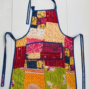 May include: A patchwork apron with a vibrant mix of colors including red, yellow, pink, and green. The apron features a blue neck strap and ties, and is made from various fabric patterns. The design includes a pocket and a quilted texture.