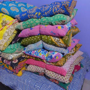 May include: A stack of colorful, patterned pillows in various colors including blue, pink, green, and yellow. The pillows have a quilted texture and are arranged on a blue and white striped surface.