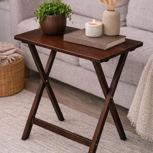 May include: A dark brown, folding side table with an X-shaped base. The tabletop holds a potted plant, a lit candle, a book, and a small vase with dried flowers. The table is made of wood and sits on a light gray rug.