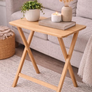 May include: A small, light-brown folding side table with an X-frame base. The tabletop holds a potted plant, a candle, a book, and a decorative vase. The table is set on a patterned rug in front of a light-colored sofa.