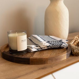 May include: A wooden serving tray with a candle in a glass jar, a stack of striped napkins, and a cream-colored vase. The candle has a label that reads "White Jasmine Aromatic Candle". The napkins have blue stripes.