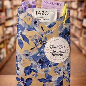 May include: A gift-wrapped book with blue floral paper, a Tazo tea packet, a book review card, and a yellow pen. The package is labeled "Blind Date With a Book Romance" and sits on a small wooden easel.