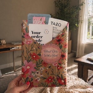 May include: A rectangular package wrapped in brown paper with a colorful floral print. A circular sticker reads "Tina's Book Date Blind Date With a Book". Inside are a 2026 Reading List card, a "Your order made my day" note, and a Tazo tea bag.