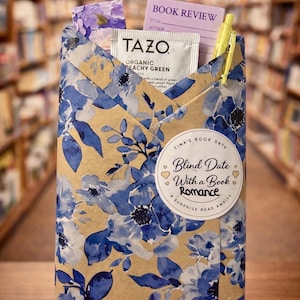 May include: A gift-wrapped book with blue floral paper, a Tazo tea packet, a book review card, and a yellow pen. The package is labeled "Blind Date With a Book Romance" and sits on a small wooden easel.