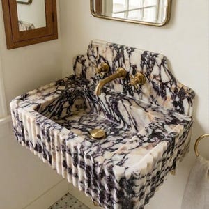 May include: A marble sink with a white and dark purple veined pattern. The sink has a fluted front and gold-coloured fixtures, including taps and a drain cover. A towel ring is mounted on the wall.