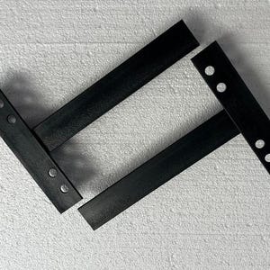 K&ouml;nnte beinhalten: Zwei schwarze Regalhalterungen aus Metall, jeweils mit einem langen horizontalen Arm und einer vertikalen Montageplatte. Die vertikalen Platten sind mit jeweils zwei kreisf&ouml;rmigen L&ouml;chern zur Befestigung versehen und bieten eine stabile St&uuml;tzstruktur.