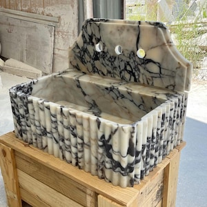 May include: A marble sink with a rectangular basin and a decorative back. The sink is white with black veining and has a fluted edge. It sits on a wooden crate. The sink has three faucet holes.