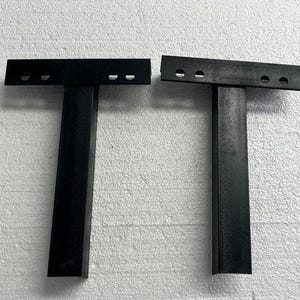 K&ouml;nnte beinhalten: Zwei schwarze, T-f&ouml;rmige Metallhalterungen, die jeweils aus einem vertikalen, rechteckigen Pfosten bestehen, der an einer horizontalen Kopfplatte befestigt ist. Die Kopfplatte jeder Halterung hat zwei kreisf&ouml;rmige L&ouml;cher zur Montage.