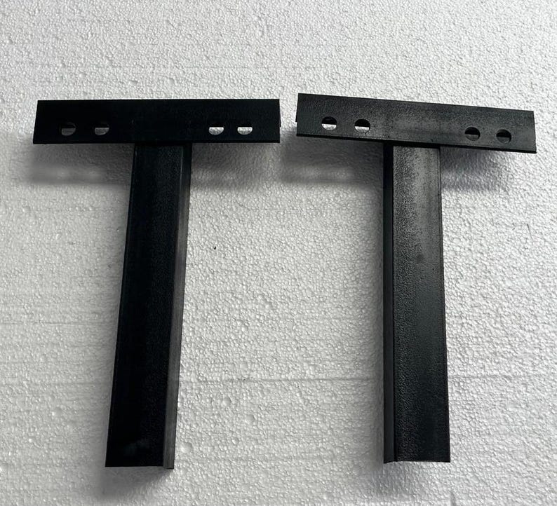 K&ouml;nnte beinhalten: Zwei schwarze, T-f&ouml;rmige Metallhalterungen, die jeweils aus einem vertikalen, rechteckigen Pfosten bestehen, der an einer horizontalen Kopfplatte befestigt ist. Die Kopfplatte jeder Halterung hat zwei kreisf&ouml;rmige L&ouml;cher zur Montage.