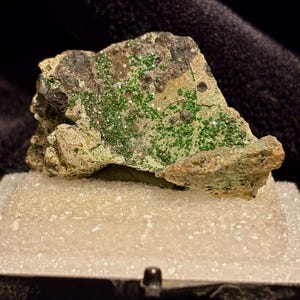 May include: A close-up of a mineral specimen featuring a rough, textured rock with patches of vibrant green crystals. The rock has a mix of brown, gray, and tan hues, and is displayed on a white, textured surface.