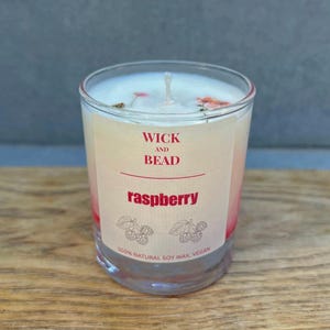 May include: A clear glass candle with a white wax top and a wick. The candle has a label that reads "WICK AND BEAD raspberry" with illustrations of raspberries. The candle is made with 100% natural soy wax and is vegan.