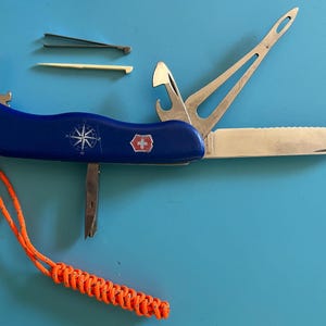 May include: A blue multi-tool with a compass rose and cross emblem. The tool includes a knife, bottle opener, and other implements. An orange lanyard is attached. Additional tools, including tweezers and a toothpick, are also visible.