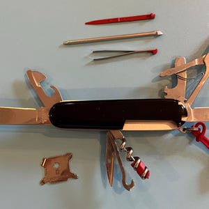 May include: A multi-tool with a black handle and various stainless steel implements, including a knife, scissors, and bottle opener. Additional tools include a toothpick, tweezers, and a corkscrew. A red lanyard is attached. The tools are displayed open.